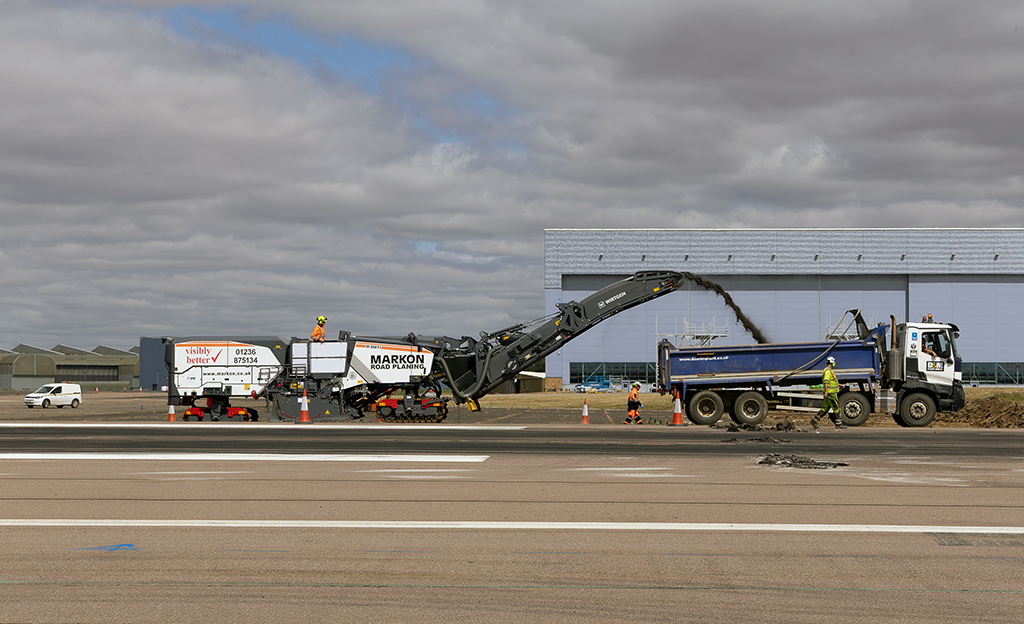 A large Markon Road Planning machine extracts asphalt from the runway and deposits it into the back of a blue and white tipper lorry on the runway. Construction workers in high-visibility clothing stand nearby under a cloudy sky, with a large blue hangar visible in the background.